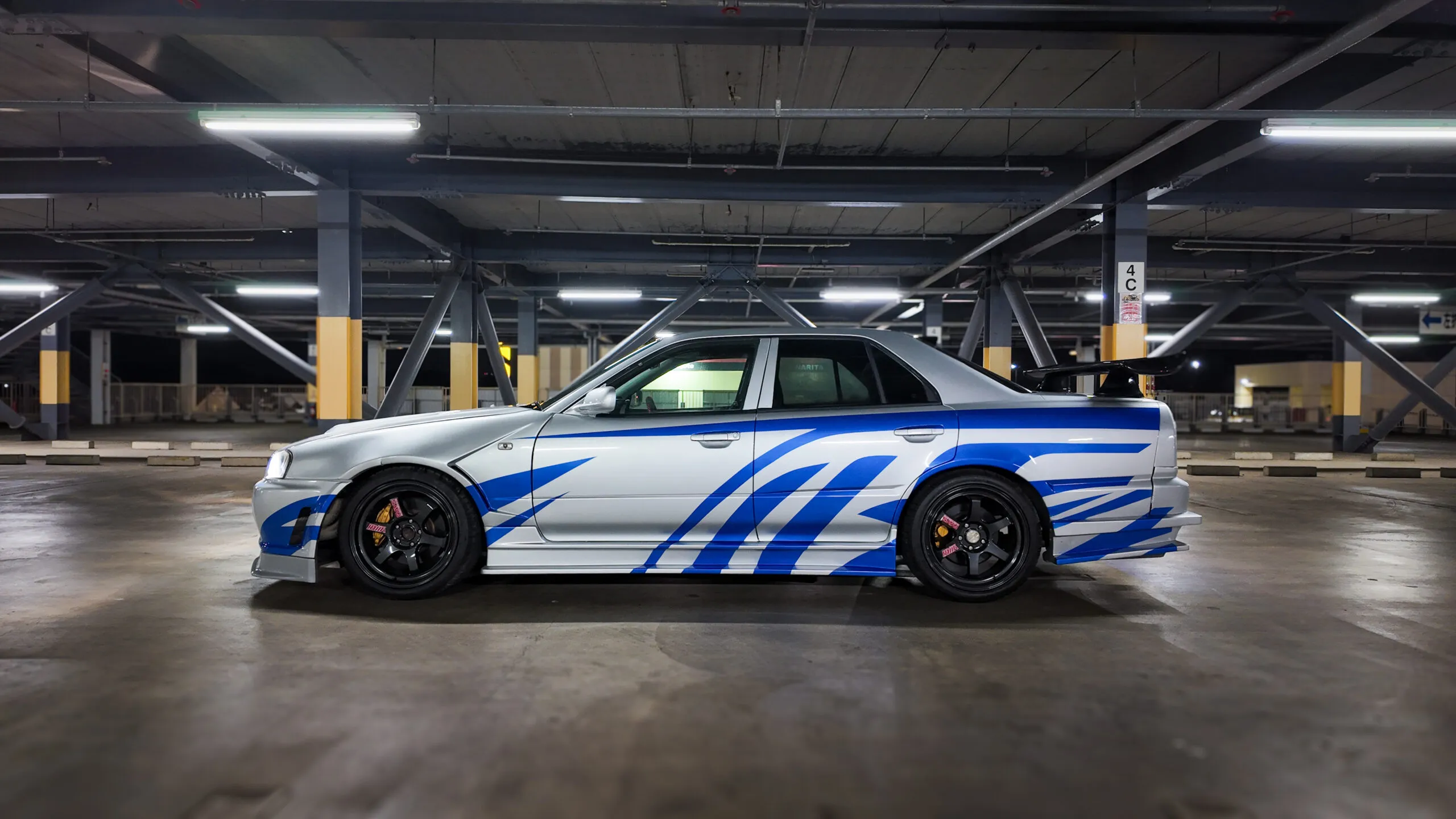 Side profile of a Nissan Skyline GT-R R34 with blue decals, black wheels, and lowered suspension, parked under fluorescent lights.